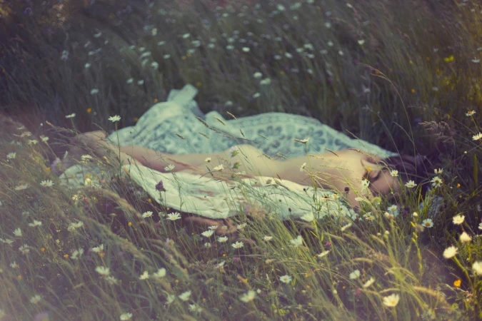 Young woman lies in a meadow of daisies in soft light, wearing a white dress and blanket, with an eye-level, outdoor view.