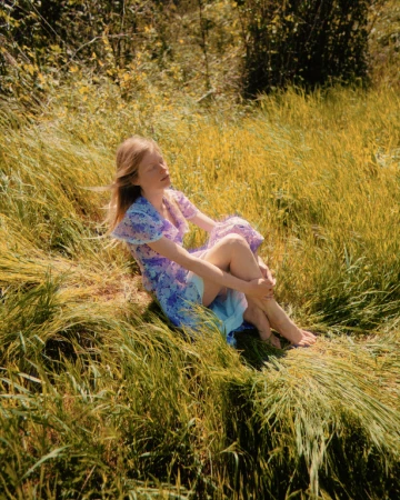 Woman in a blue floral dress seated in tall golden grass with eyes closed