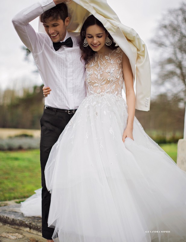 A smiling couple stands close together outdoors, holding a white jacket overhead like a canopy. The woman wears a white bridal gown.