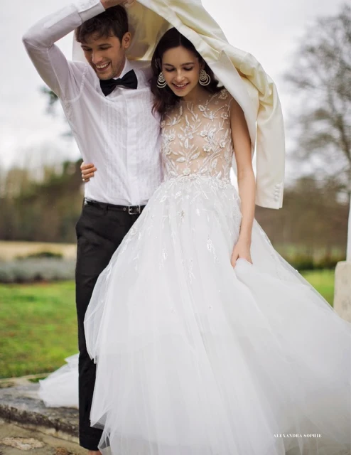 A smiling couple stands close together outdoors, holding a white jacket overhead like a canopy. The woman wears a white bridal gown.