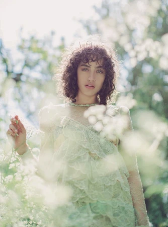 Portrait of a woman with dark curly hair wearing a pale lace dress surrounded by out of focus white wildflowers.