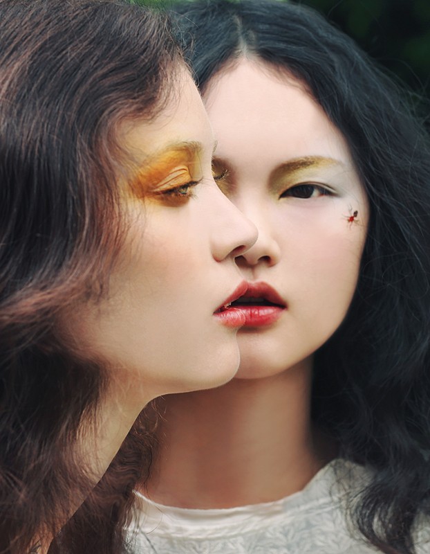 Close-up portrait of two Asian women with dramatic makeup, one with golden eye shadow, the other with red lips, in a blurred outdoor setting.