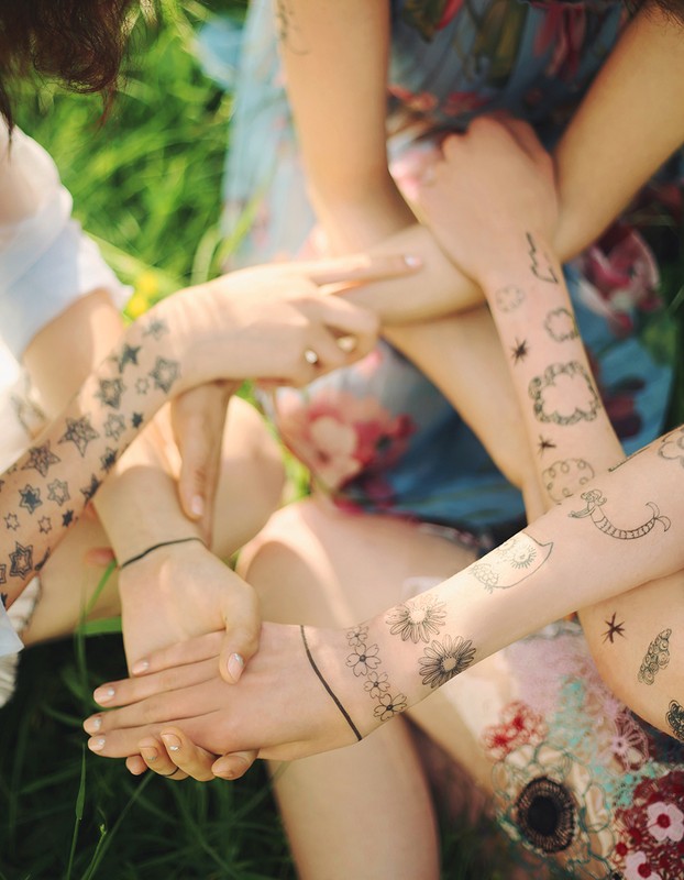 Close-up of arms and hands of three young women in an outdoor setting, showing temporary tattoos and bracelets with a soft focus.