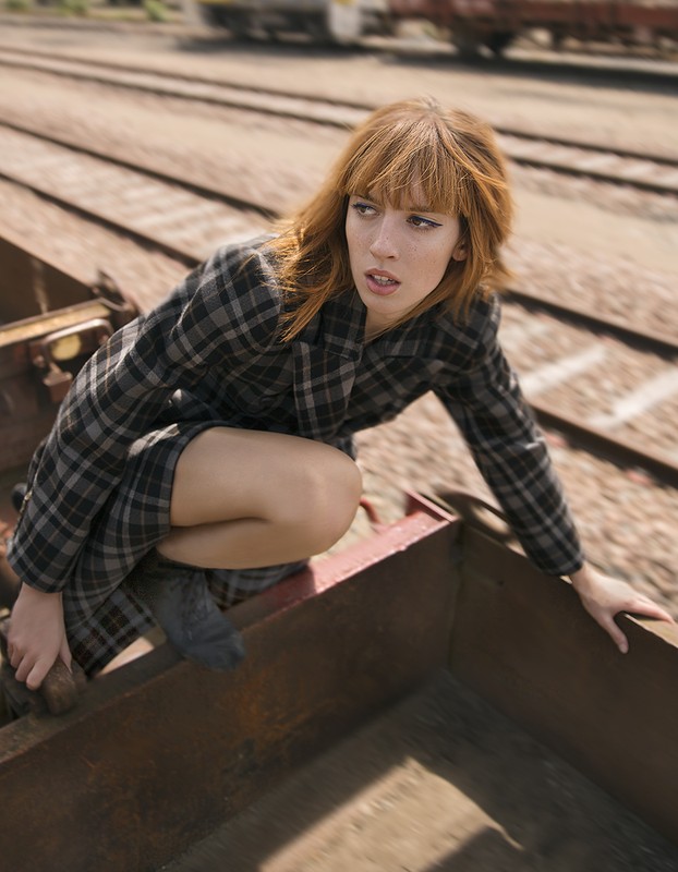 Woman in a checkered coat crouches on a train car near train tracks, looking off to the side with a serious expression in daylight.