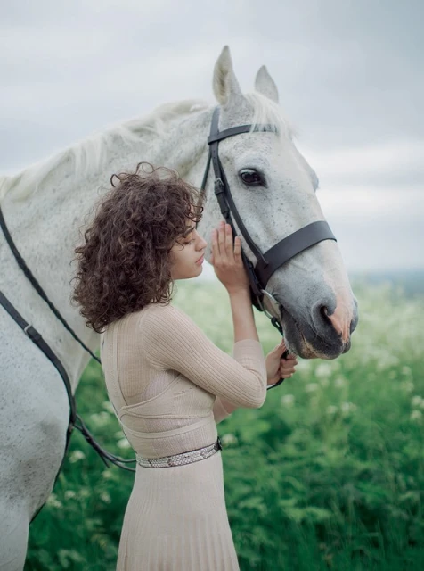 Young woman with curly hair gently touches the muzzle of a white horse in a field of wildflowers, eye-level shot, natural light.