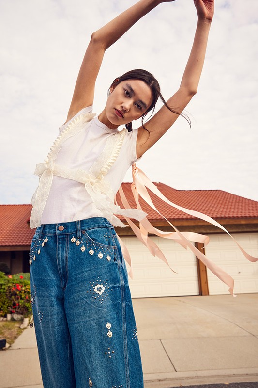 Woman in denim jeans poses outdoors wearing a white tee, lace harness, and ribbons against a backdrop of a suburban home on a sunny day.