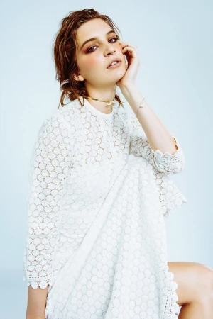A woman wearing a white lace dress with her hand to her cheek against a pale background, styled by Alison Lewis.