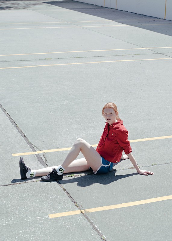 Redheaded woman seated on an asphalt parking lot wearing a red jacket and blue shorts styled by Alison Lewis.