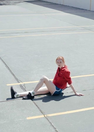 Redheaded woman seated on an asphalt parking lot wearing a red jacket and blue shorts styled by Alison Lewis.