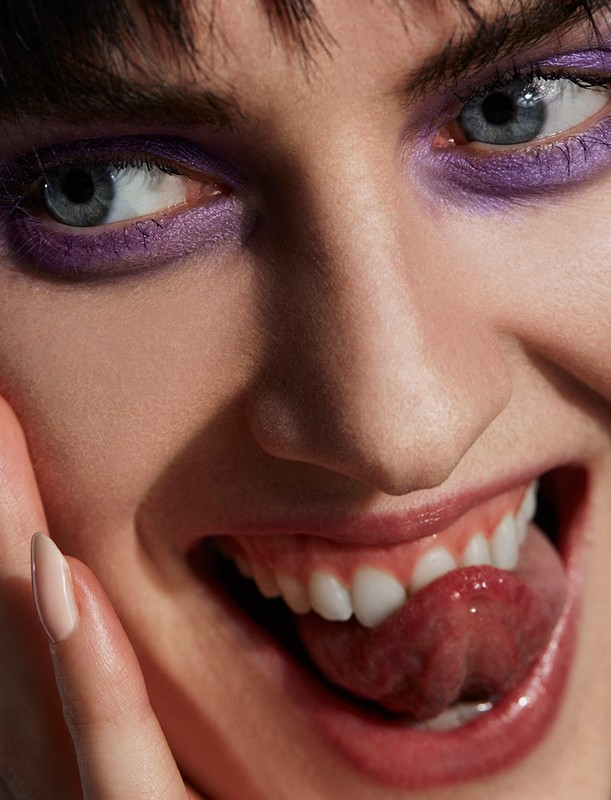 Close-up portrait of a woman with vibrant purple eye makeup sticking out her tongue styled by Alison Lewis