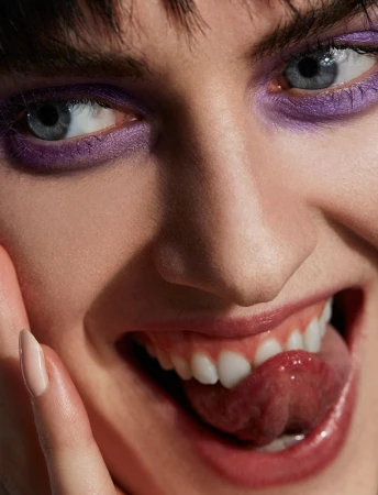 Close-up portrait of a woman with vibrant purple eye makeup sticking out her tongue styled by Alison Lewis