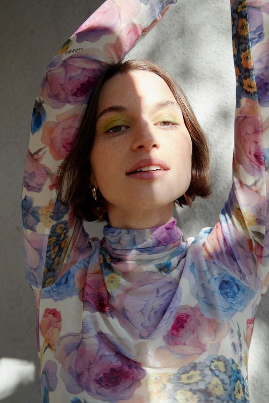 Close-up portrait of a woman with brown hair and green eye shadow, wearing a floral print top, posing in front of a gray background.