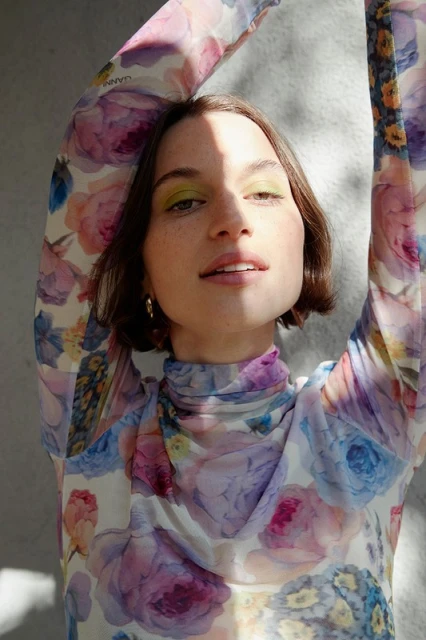 Close-up portrait of a woman with brown hair and green eye shadow, wearing a floral print top, posing in front of a gray background.