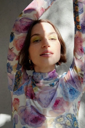 Close-up portrait of a woman with brown hair and green eye shadow, wearing a floral print top, posing in front of a gray background.