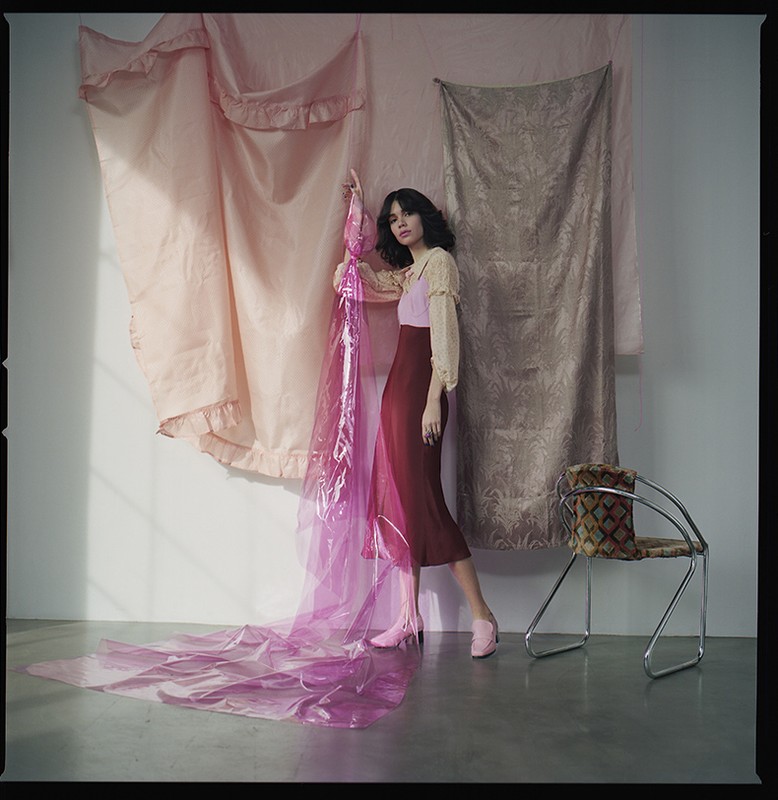 Woman in a burgundy dress poses in a studio with draped pink fabric, complemented by a chair with patterned upholstery.