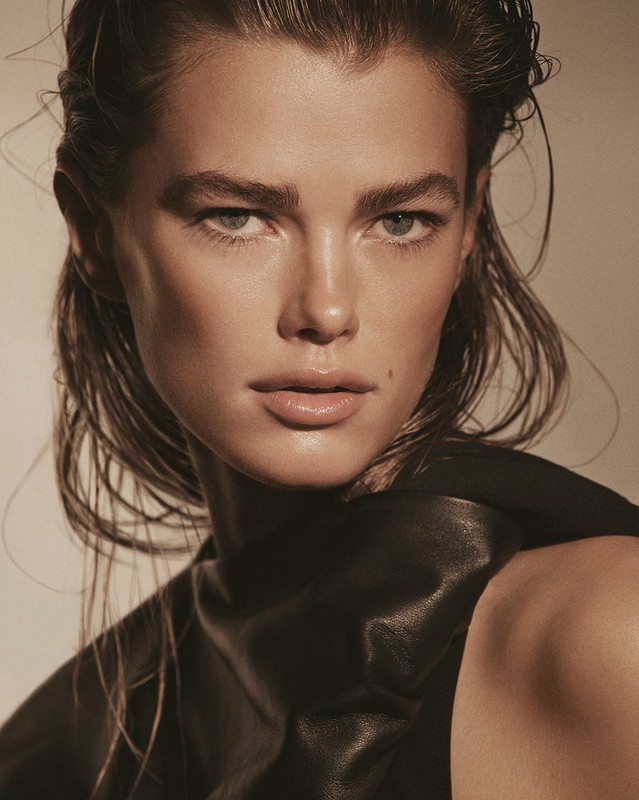 Close-up portrait of an adult female fashion model with wet hair, wearing a black leather top, set against a neutral-toned backdrop.