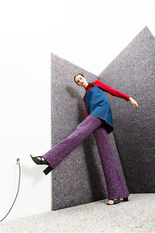 A fashion shot of a woman in a red top, denim dress, and purple pants posing indoors against a grey textured background with an outlet.
