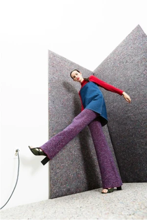 A fashion shot of a woman in a red top, denim dress, and purple pants posing indoors against a grey textured background with an outlet.