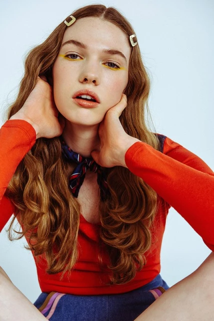 Young woman in red top and hair clips with wavy brown hair poses against a light background, looking directly at the camera.