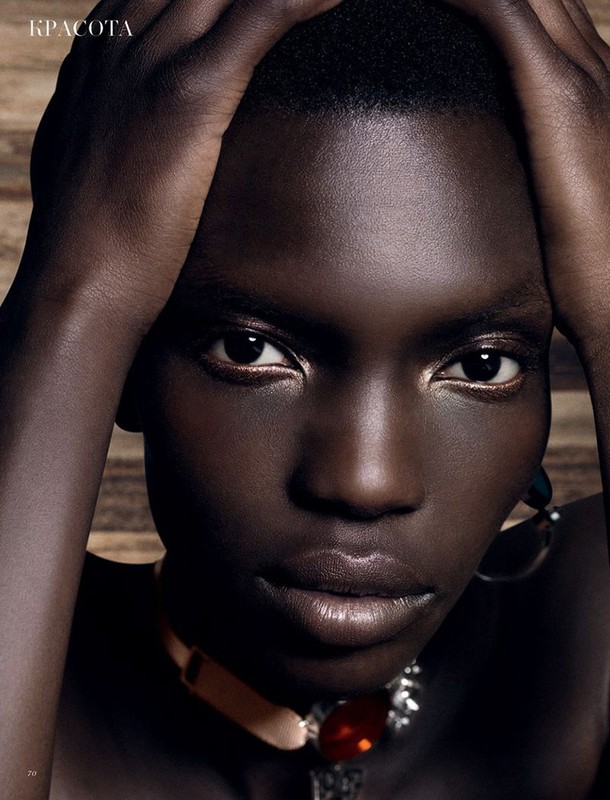 Close-up portrait of a dark-skinned woman with hands near her face, adorned with statement jewelry, against a blurred wooden background.