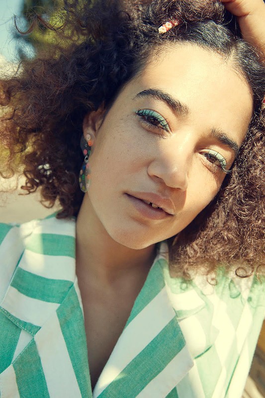 Close-up portrait of a smiling woman with curly hair wearing a striped blazer outdoors on a sunny day, showcasing fashion and beauty.