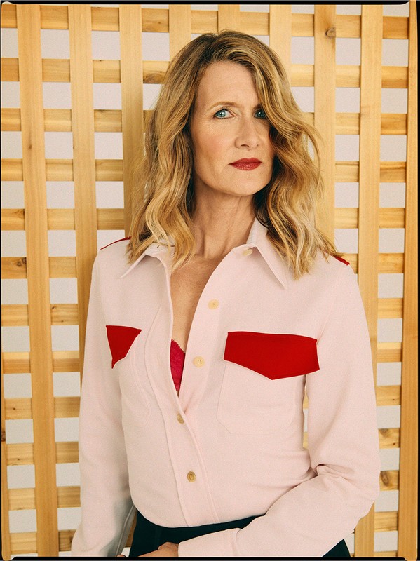 Portrait of Laura Dern wearing a pink collared shirt with red accents, posing in a photo studio with a natural wood backdrop.