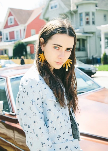 Portrait of a young woman with long brown hair and gold flower earrings standing near a vintage car and houses in a residential area.