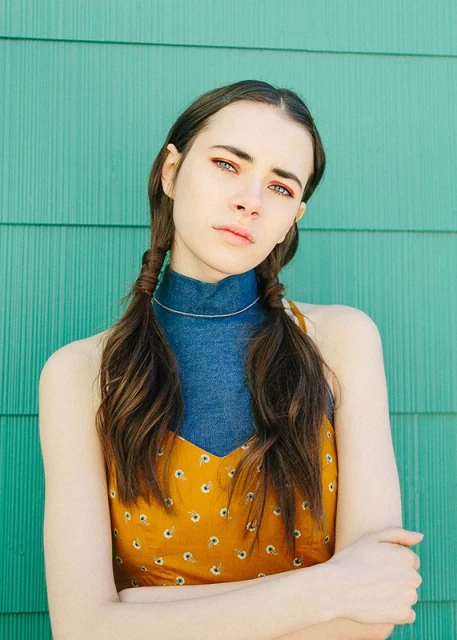 Portrait of a young woman with long brown hair in pigtails wearing a floral dress in front of a turquoise-painted wall, arms crossed.