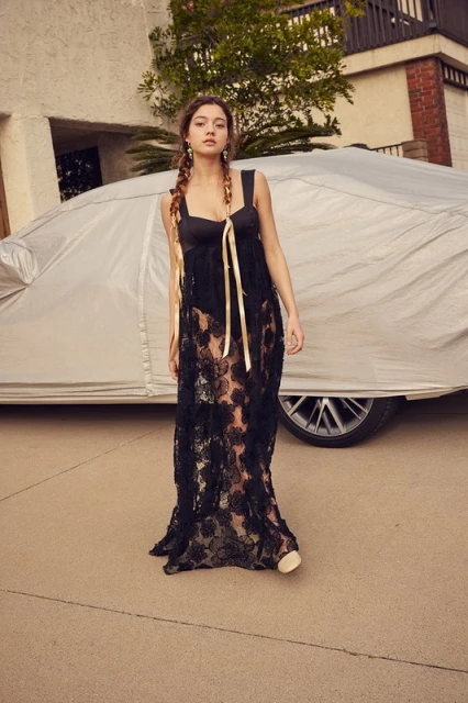 A full-length portrait of a young woman in a black lace dress with a ribbon tie, standing in front of a car covered with a silver tarp.