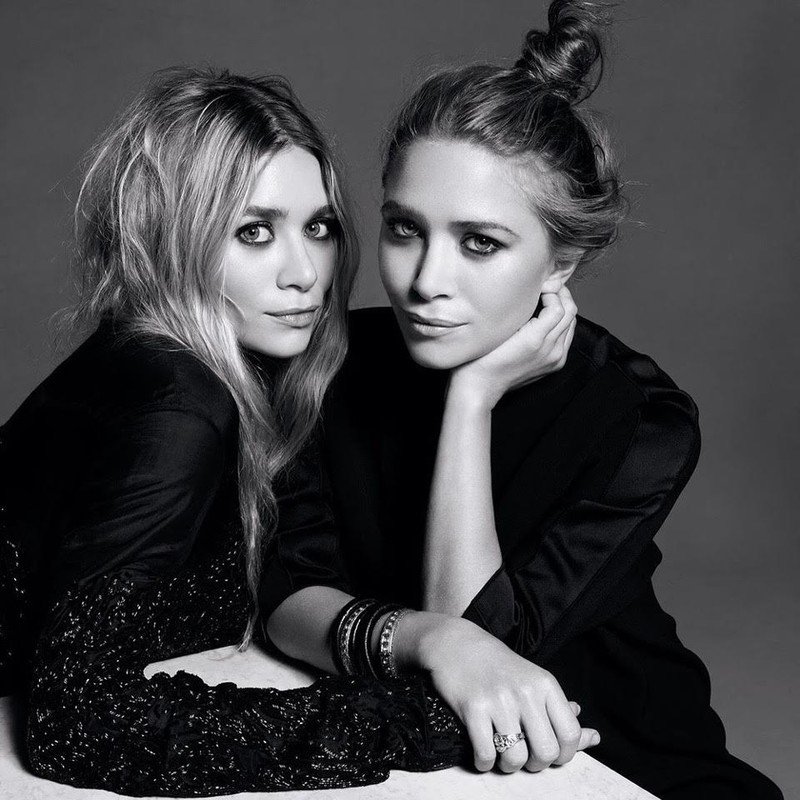Black and white portrait of two adult women with dark eye makeup wearing dark, silky tops, posing together in a photo studio setting.