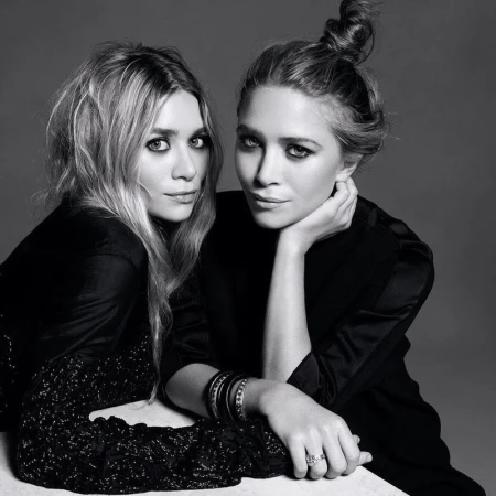 Black and white portrait of two adult women with dark eye makeup wearing dark, silky tops, posing together in a photo studio setting.