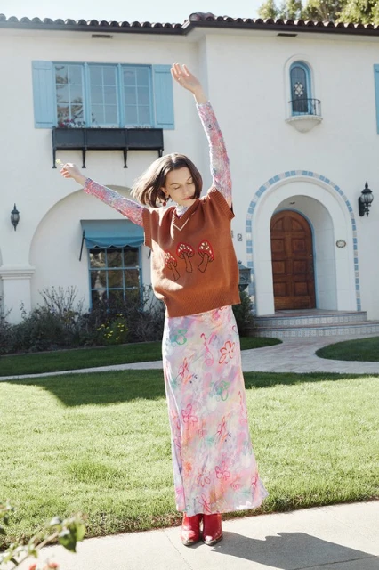 Adult female in mushroom sweater and floral skirt smiles in front of a two-story white house with lawn and plants on a sunny day.