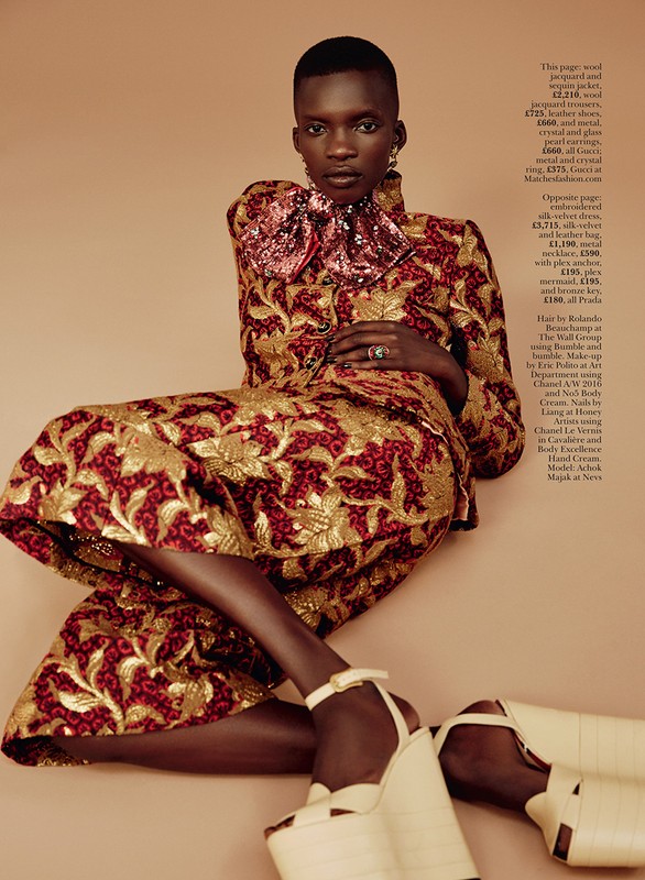 Black woman with short hair wearing a patterned metallic jacquard jacket and matching skirt, seated against a solid background.