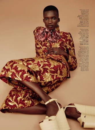 Black woman with short hair wearing a patterned metallic jacquard jacket and matching skirt, seated against a solid background.