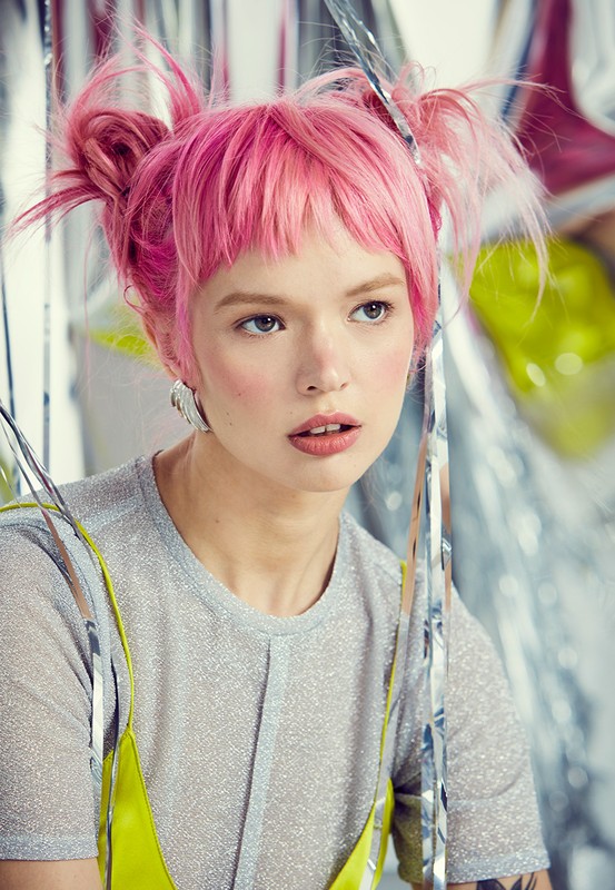 Portrait of a woman with vibrant pink space buns, wearing a silver shirt with a chartreuse top, and set against a shiny silver background.