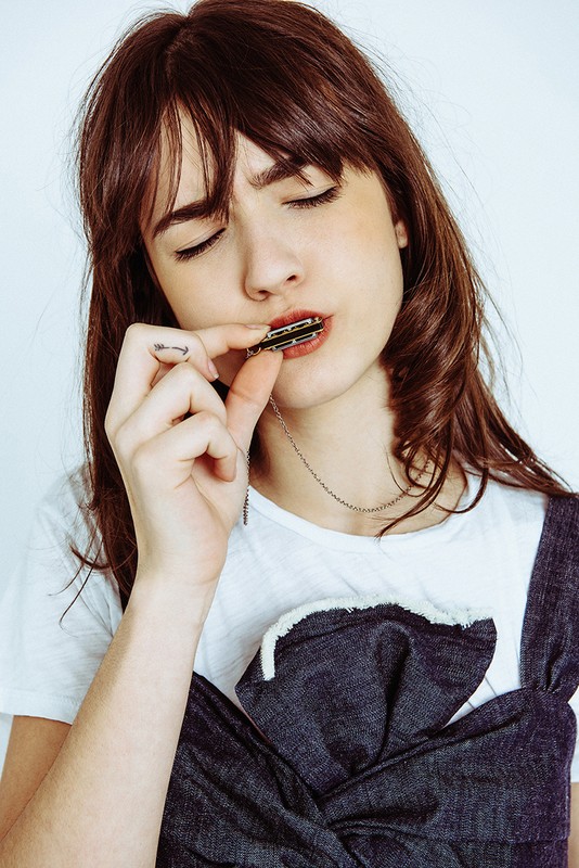 Young woman with dark hair and bangs wearing a t-shirt with denim overall dress holding a harmonica with eyes closed in a studio setting.