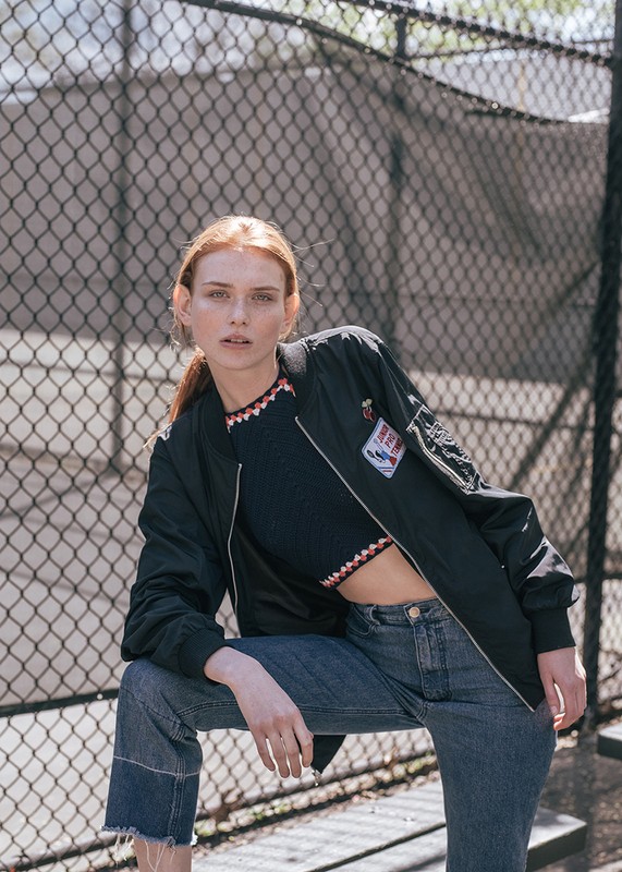 A young red-haired woman in a sporty outfit poses in front of a chainlink fence with concrete pillars in the background on a sunny day.