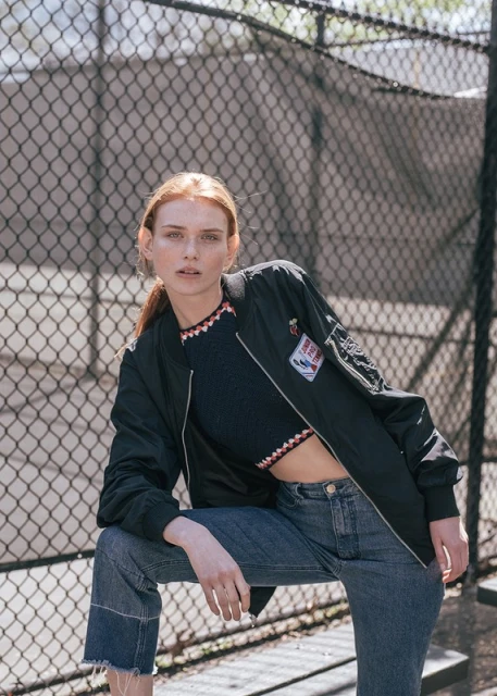 A young red-haired woman in a sporty outfit poses in front of a chainlink fence with concrete pillars in the background on a sunny day.