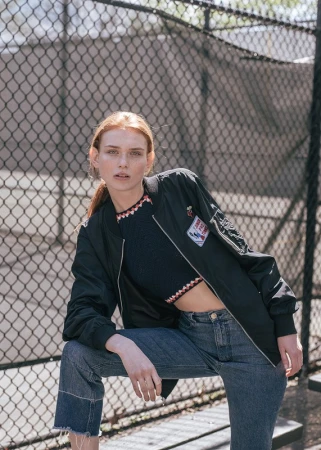 A young red-haired woman in a sporty outfit poses in front of a chainlink fence with concrete pillars in the background on a sunny day.