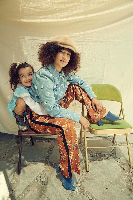 Adult and child fashion portrait. An adult female with curly hair wears a hat and floral pants while a child sits on a chair behind her.