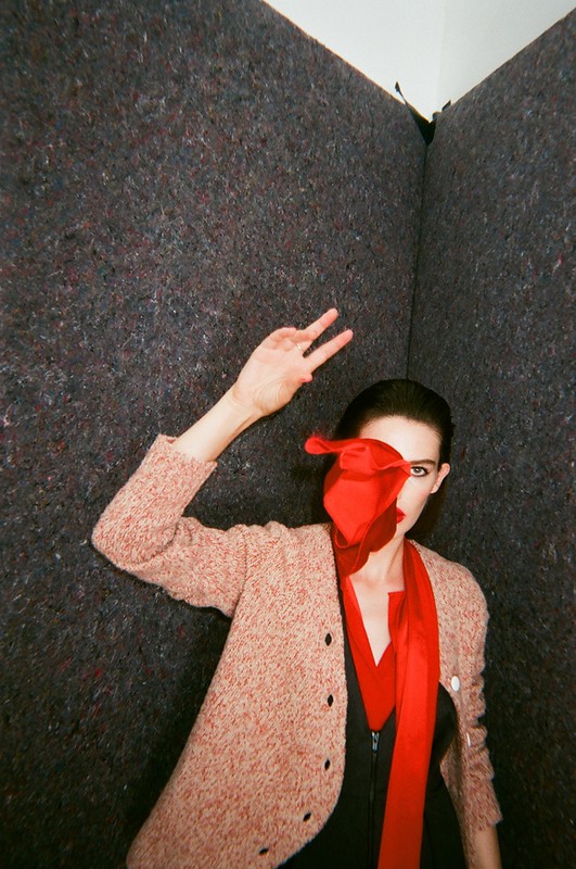Fashion portrait of a woman in a pink sweater and red scarf, striking a pose with a two-finger gesture in a textured grey corner.