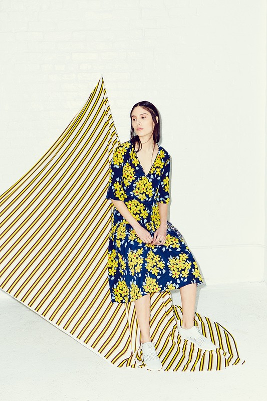 Brunette woman poses in a blue wrap dress with yellow floral print, sitting on a striped backdrop in a white photo studio.