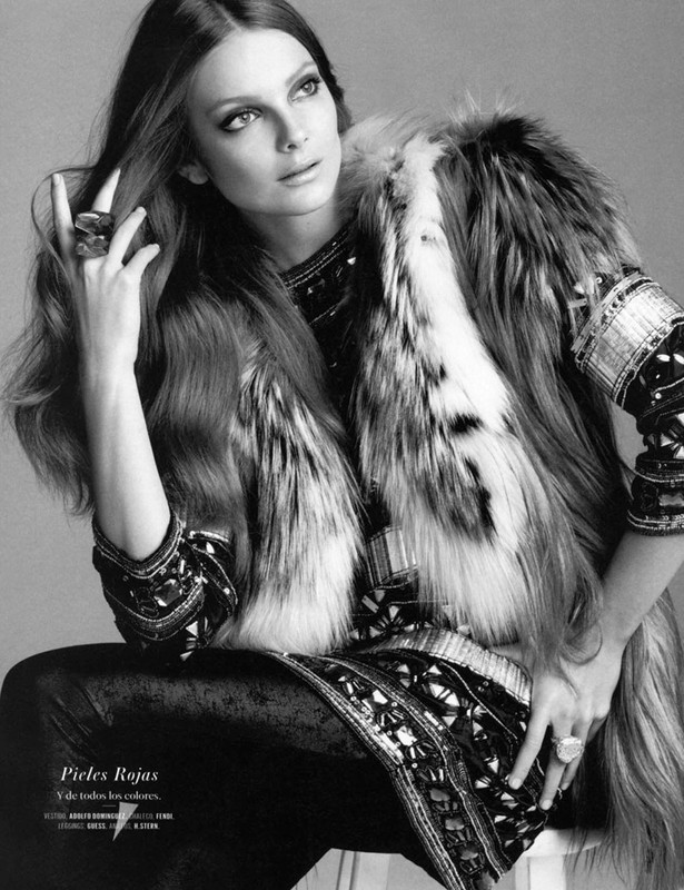 Black and white portrait of a fair-skinned adult woman with long brunette hair, wearing a fur vest and ornate top, seated indoors.