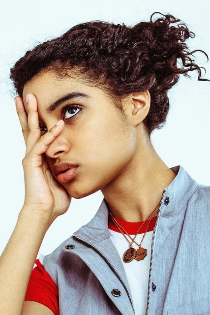 Portrait of a curly-haired young woman with hand near face, wearing a sleeveless gray vest over red shirt against a plain white background.