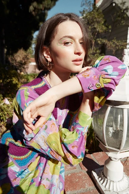 A woman with short brown hair in a patterned dress poses next to a lamp post outside on a sunny day in an eye-level medium shot.