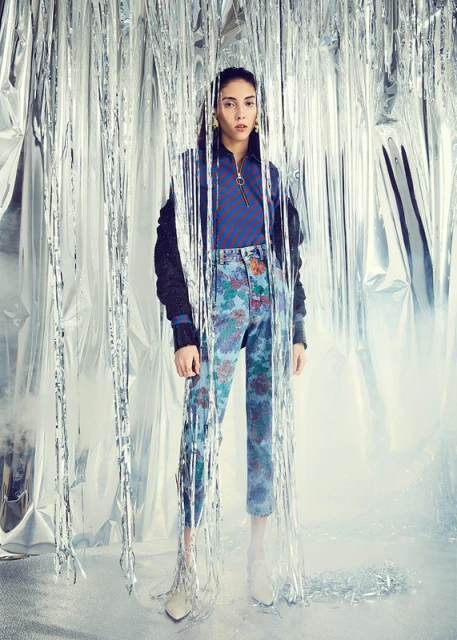 Fashion shot of a woman in floral pants and a bomber jacket standing in front of a silver tinsel backdrop in a studio setting.
