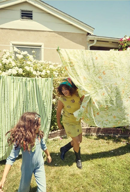 Two females in retro clothing playfully interact in a sunlit backyard with vibrant green lawn and flowering bushes creating a nostalgic scene.