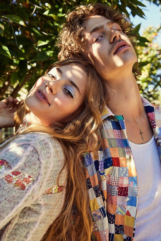 A young couple outdoors in sunlight, the woman rests her head on the man, both are wearing patterned clothing and looking at the camera.