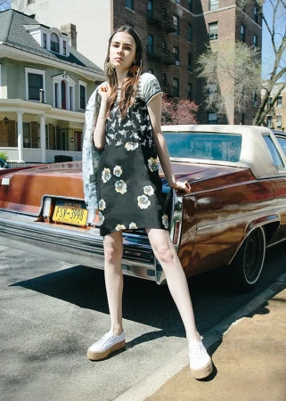 Young woman standing in front of a vintage brown car wearing a black floral dress and white platform sneakers