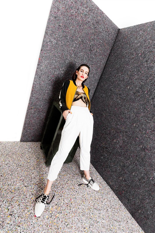 A woman poses in a fashion shot with a yellow and black bomber jacket, crop top, and white pants in front of a gray textured backdrop.